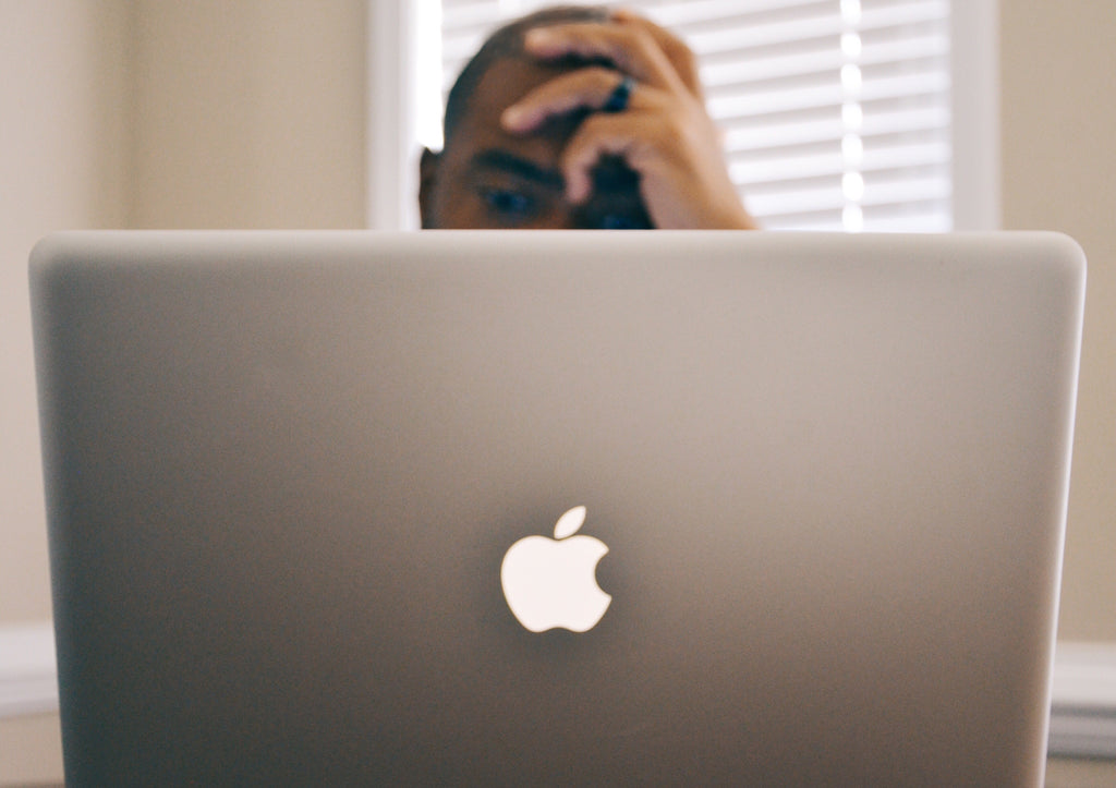 african american man with hand on forehead staring at an Apple computer in the foreground, confused and frustrated